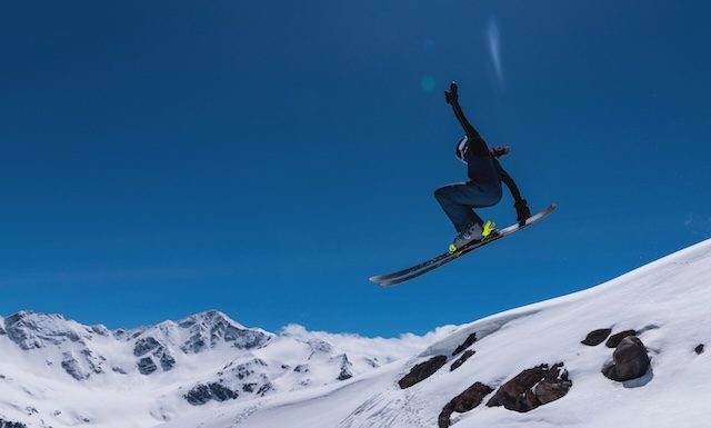 Young female skier jumping from ski jump on sunny day in snowy alpine mountains, moment in flight.