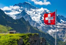 Suisse | Hiver record pour l’hôtellerie suisse : +18,5 millions de nuitées, un sommet historique Swiss flag wavingon a Mannlichen viewpoint with peak of Jungfrau mountain on the background, Bernese Oberland Switzerland @ credit Depositphotos