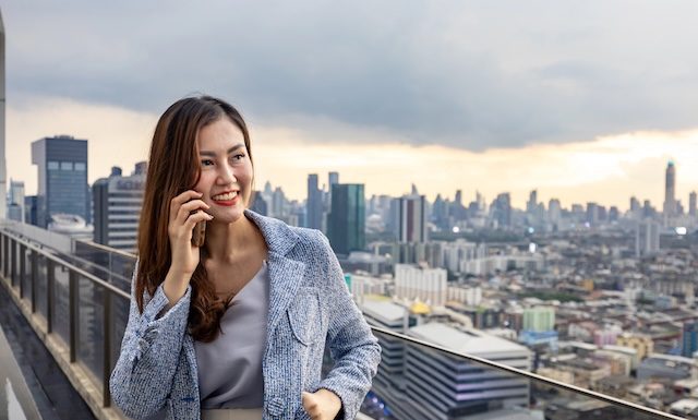 Asian business woman in formal suit is phone calling customer while standing outside skyscraper building for marketing, connection, corporate work, real estate, housing and urban development