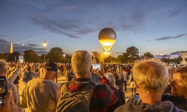 Paris, France - 07 28 2025: People standing from behind take photos with cellphones of the Olympic Cauldron as it takes flight from the Tuileries Garden at sunset.