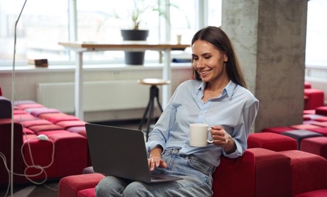 Cheerful young woman with teacup in hand looking at computer monitor while sitting on chair in comfortable coworking space