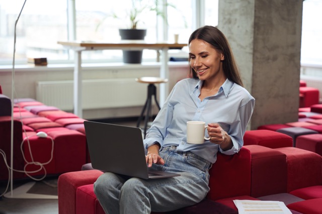 Cheerful young woman with teacup in hand looking at computer monitor while sitting on chair in comfortable coworking space