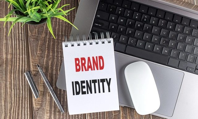 Laptop, mouse and notebook notes with text BRAND IDENTITY on wooden background.