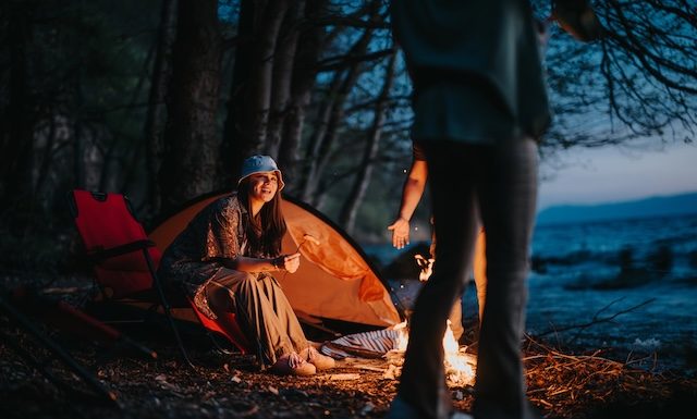 A happy camper sitting by a warm campfire near a tent and chair beside a tranquil lake as night falls.