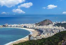 An aerial panoramic view of Copacabana beach, Rio de Janeiro, with its dense city skyline, iconic mountains, and blue Atlantic Ocean under a sunny sky @ credit Depositphotos.com