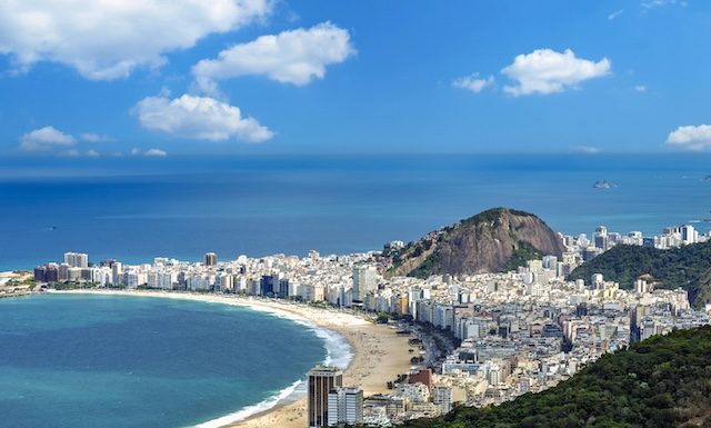 An aerial panoramic view of Copacabana beach, Rio de Janeiro, with its dense city skyline, iconic mountains, and blue Atlantic Ocean under a sunny sky @ credit Depositphotos.com